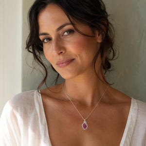 May include: A silver necklace featuring a wire-wrapped pendant with a deep purple, faceted gemstone. The pendant hangs from a delicate silver chain, resting against a white fabric garment. The design showcases intricate metalwork around the stone.