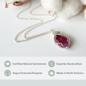 May include: A silver chain necklace featuring a wire-wrapped, irregular, deep pink gemstone pendant. Below the jewelry, text reads: Certified Natural Gemstones, Expertly Handcrafted, Vogue Featured Designer, and Made in North America.