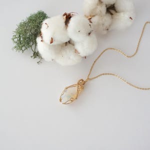 May include: A gold-toned wire-wrapped pendant necklace featuring an oval-shaped, semi-translucent white stone with subtle grey inclusions. The pendant hangs from a delicate gold-toned chain, positioned near white cotton bolls and green moss.