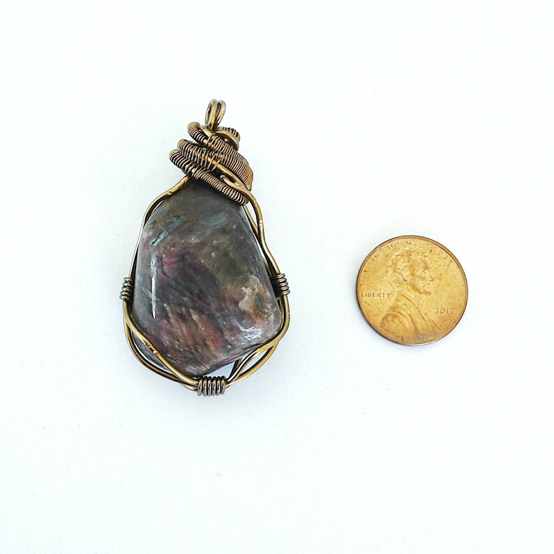 May include: A handcrafted pendant featuring a polished, multi-colored stone wrapped in bronze-colored wire. The stone displays shades of gray, pink, and green. A penny is next to the pendant for size reference.
