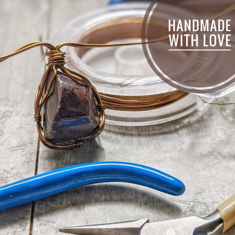 May include: A handmade pendant featuring a dark, irregularly shaped stone wrapped in copper wire. The wire is intricately woven around the stone, creating a cage-like design. The image also shows spools of copper wire, pliers, and the text "HANDMADE WITH LOVE".