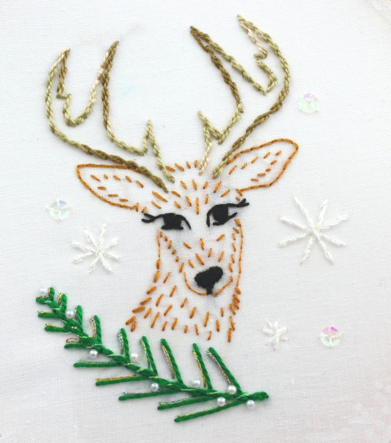 May include: Embroidered design of a brown reindeer with antlers, looking to the right, with a green pine branch with white beads underneath. The background is white with white snowflakes.