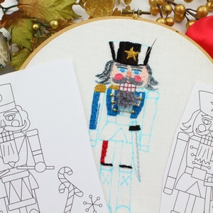 May include: A close-up of a Nutcracker embroidery project. The embroidery is on white fabric and is surrounded by a wooden embroidery hoop. The Nutcracker is wearing a blue and red uniform with a gold star on his hat. There are two black and white line drawings of Nutcrackers on paper next to the embroidery.