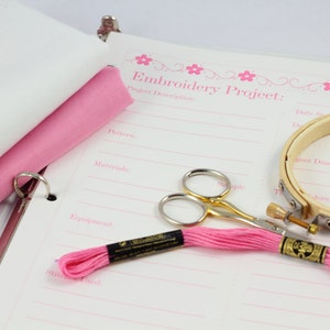 May include: A white binder with a pink fabric sample and a page titled "Embroidery Project: Project Description". The page has lines for writing down details about the project, such as the pattern, materials, equipment, and stitch used. There is also a pink embroidery floss, a pair of small gold scissors, and a wooden embroidery hoop.