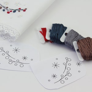 May include: Two white paper patterns with a simple design of a branch with berries and snowflakes.  There are six spools of embroidery floss in red, blue, gray, and brown.