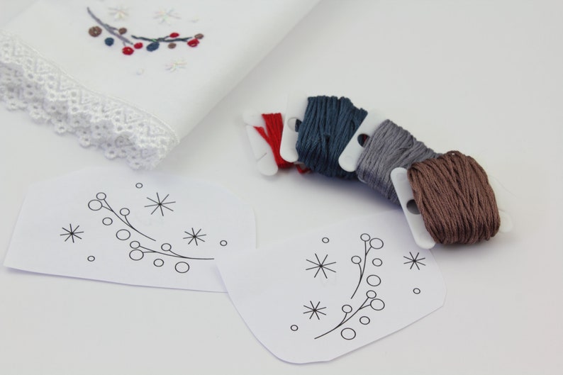 May include: Two white paper patterns with a simple design of a branch with berries and snowflakes.  There are six spools of embroidery floss in red, blue, gray, and brown.
