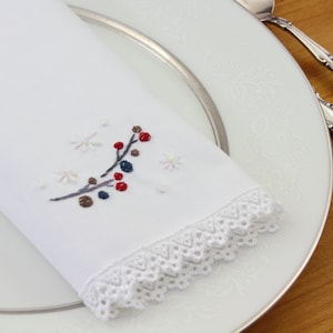 May include: White linen napkin with a delicate lace trim and embroidered winter scene featuring a branch with red and blue berries and snowflakes.