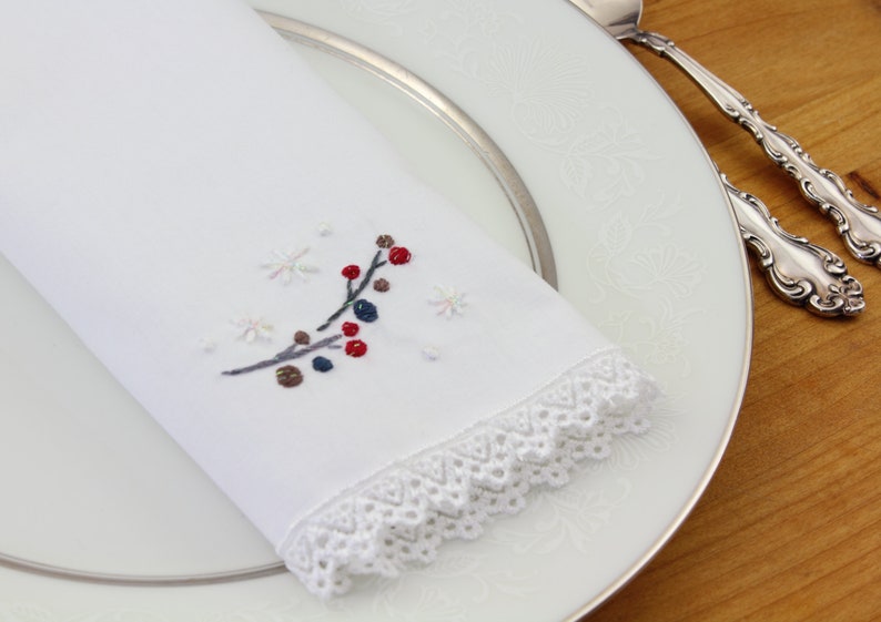 May include: White linen napkin with a delicate lace trim and embroidered winter scene featuring a branch with red and blue berries and snowflakes.