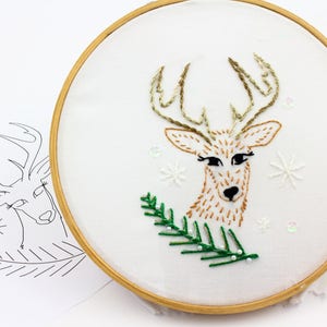 May include: Embroidered hoop art featuring a brown deer with green pine branches and white snowflakes. The deer has black eyes and a brown nose.