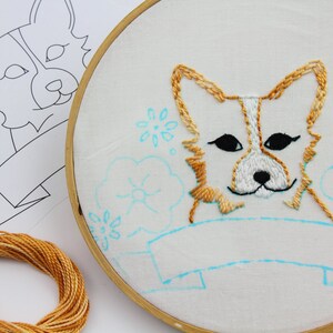 May include: Embroidered hoop art featuring a cute cartoon corgi with a blank banner below. The corgi is stitched in shades of brown and white, with black eyes. The banner is outlined in blue, and the background is filled with blue flowers.