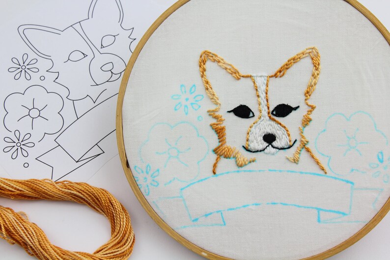 May include: Embroidered hoop art featuring a cute cartoon corgi with a blank banner below. The corgi is stitched in shades of brown and white, with black eyes. The banner is outlined in blue, and the background is filled with blue flowers.