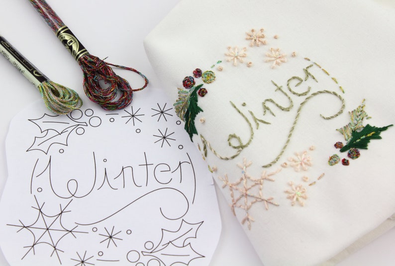 May include: A white fabric with green and red embroidery of the word "Winter" surrounded by snowflakes and holly leaves. A printed pattern of the same design is shown next to it with two spools of embroidery floss.