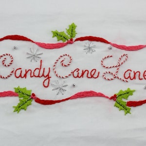 May include: Embroidered design with the words "Candy Cane Lane" in red script. The design includes red and pink ribbons, candy canes, snowflakes, and green holly leaves, all on a white background.