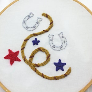 May include: Embroidered hoop art with a brown rope design, two silver horseshoes, and red and blue stars.