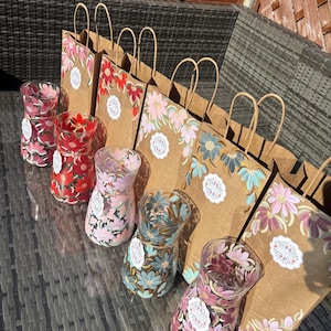 May include: A selection of hand-painted glass vases featuring floral patterns in pink, red, and blue hues. The vases are arranged in front of brown paper gift bags, similarly adorned with flowers. The image is taken in natural sunlight.
