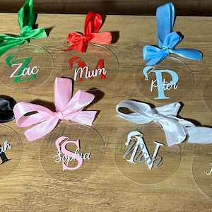 Puede incluir: Adornos redondos de acrílico transparente con nombres y lazos de cinta de varios colores. Los nombres están escritos en verde, rojo, azul, amarillo, rosa, negro y blanco. Los nombres incluyen Zac, Mum, Peter, Ashlee, Hannah, Sophia, N y Jessica.