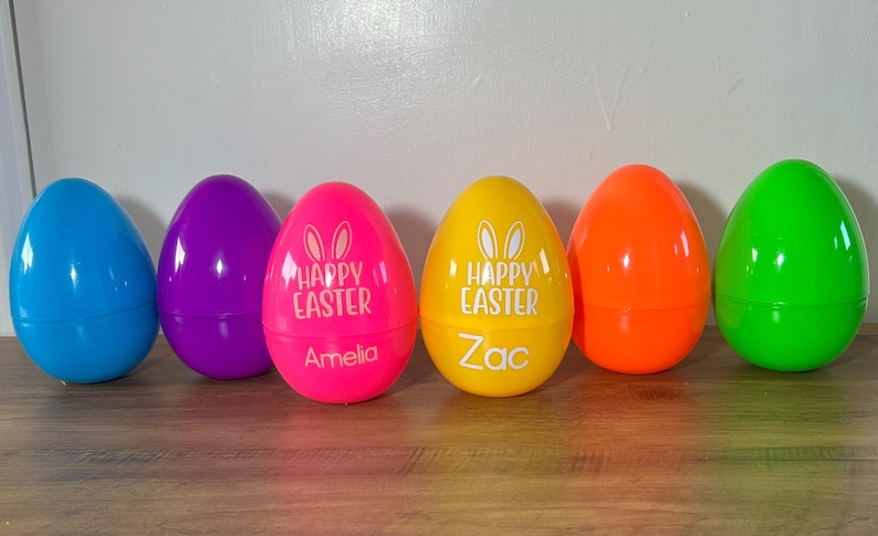 May include: Six plastic Easter eggs in various colors, including blue, purple, pink, yellow, orange, and green. The eggs are decorated with bunny ears and the words "Happy Easter". Two of the eggs are personalized with the names "Amelia" and "Zac".