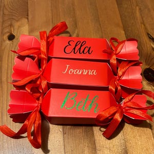 Peut inclure: Trois pétards de Noël rouges, attachés avec des rubans rouges. Chaque pétard porte un nom : "Ella" en noir, "Joanna" en or et "Beth" en vert. Ils sont posés sur une surface en bois.
