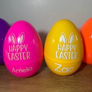 May include: Two plastic Easter eggs, one pink and one yellow, with the words "Happy Easter" and bunny ears printed on them. The pink egg has the name "Amelia" written on it, and the yellow egg has the name "Zac" written on it.