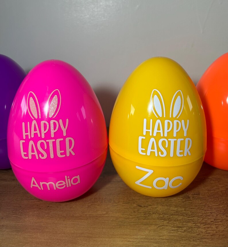 May include: Two plastic Easter eggs, one pink and one yellow, with the words "Happy Easter" and bunny ears printed on them. The pink egg has the name "Amelia" written on it, and the yellow egg has the name "Zac" written on it.