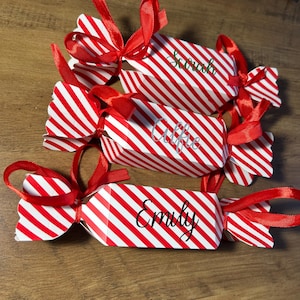 Peut inclure: Trois boîtes cadeaux en forme de bonbons, rayées rouge et blanc, nouées avec des rubans rouges. Chaque boîte porte un nom : Sarah, Alfie et Emily. Les boîtes sont disposées sur une surface en bois.