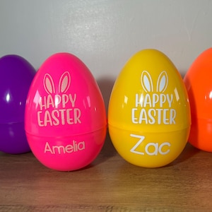 May include: Three plastic Easter eggs, one pink, one yellow, and one orange. The pink egg has "Happy Easter" and "Amelia" written on it. The yellow egg has "Happy Easter" and "Zac" written on it.