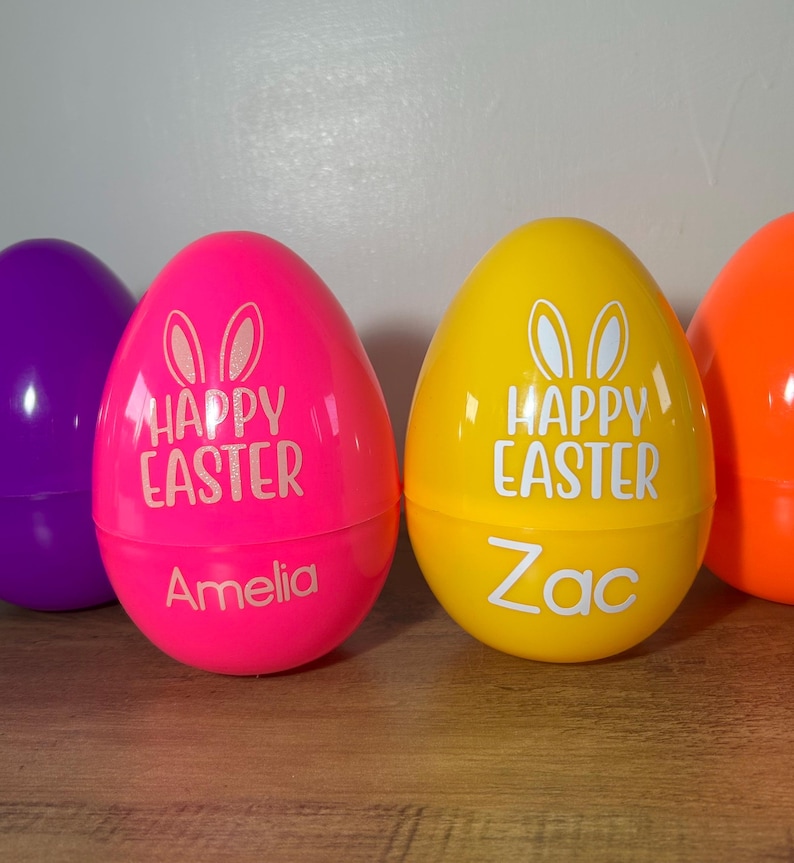 May include: Three plastic Easter eggs, one pink, one yellow, and one orange. The pink egg has "Happy Easter" and "Amelia" written on it. The yellow egg has "Happy Easter" and "Zac" written on it.