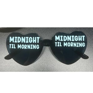 Può includere: Occhiali da sole neri a forma di cuore con la scritta bianca "MIDNIGHT TIL MORNING". Gli occhiali da sole hanno un design elegante e moderno. Le lenti sono scure e la montatura è nera.