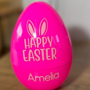 May include: Pink plastic Easter egg with bunny ears and the text "Happy Easter" and the name "Amelia" in white.