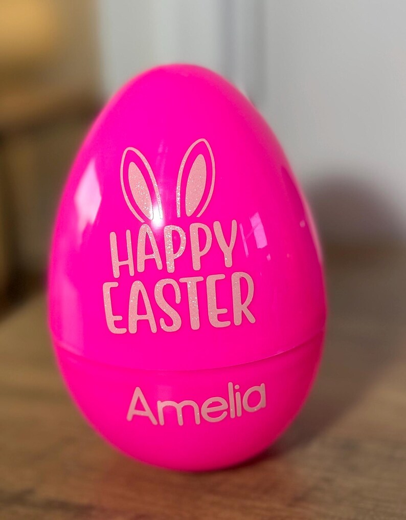May include: Pink plastic Easter egg with bunny ears and the text "Happy Easter" and the name "Amelia" in white.