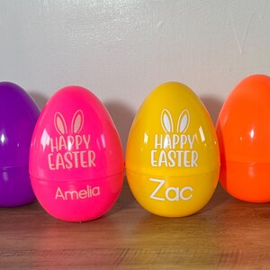 May include: Six plastic Easter eggs in various colors, including blue, purple, pink, yellow, orange, and green. The pink and yellow eggs have the text "Happy Easter" and bunny ears printed on them. The pink egg has the name "Amelia" printed on it, and the yellow egg has the name "Zac" printed on it.