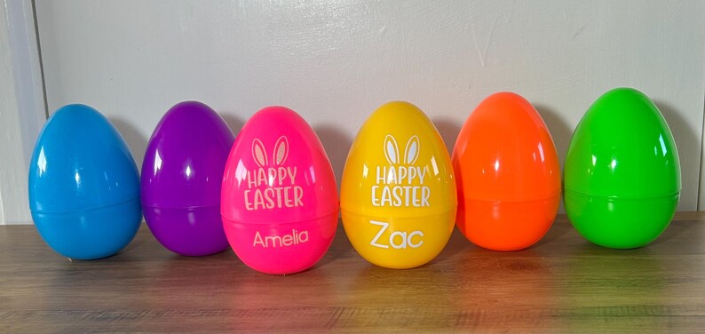 May include: Six plastic Easter eggs in various colors, including blue, purple, pink, yellow, orange, and green. The pink and yellow eggs have the text "Happy Easter" and bunny ears printed on them. The pink egg has the name "Amelia" printed on it, and the yellow egg has the name "Zac" printed on it.