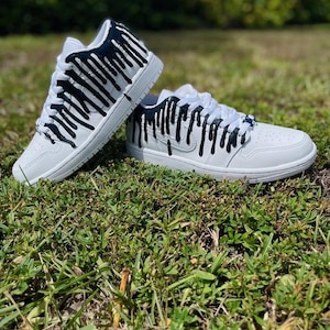 May include: White trainers with black paint dripping down the sides. The trainers are on a bed of green grass.