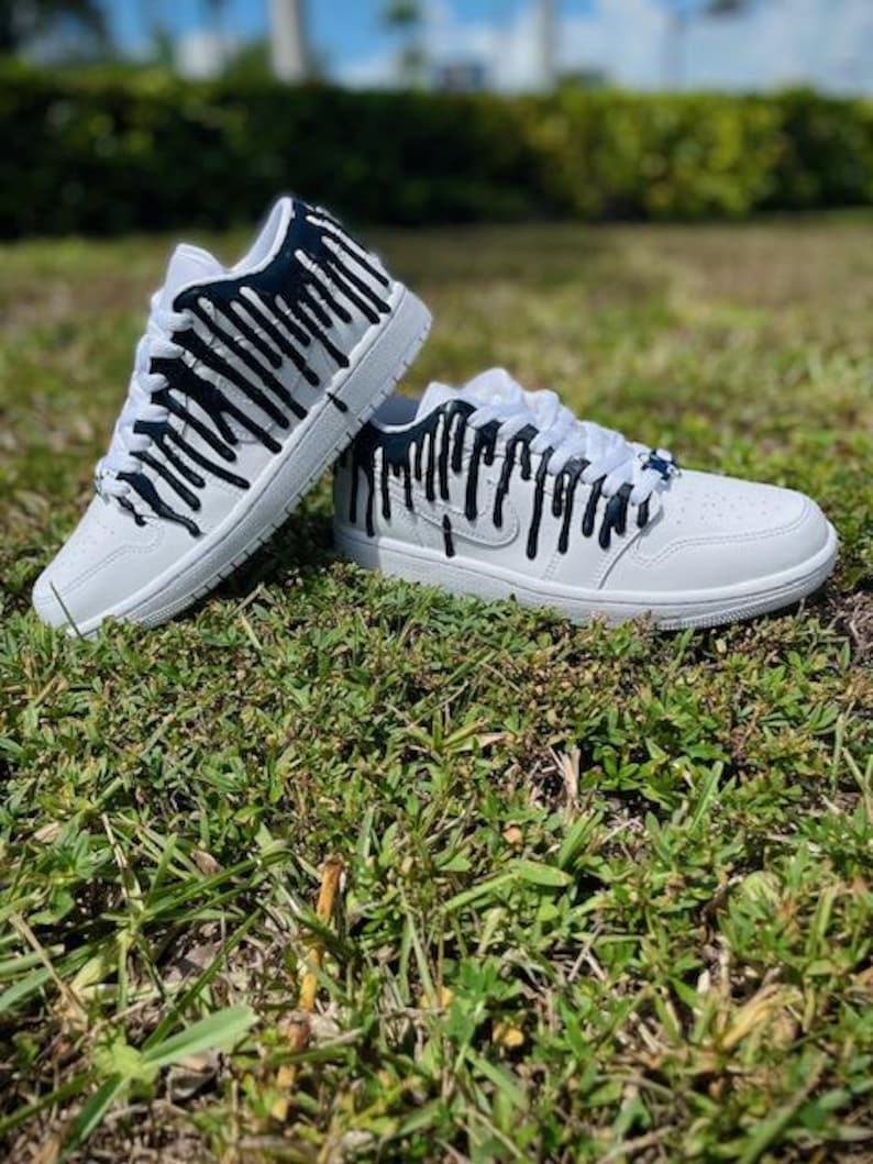 May include: White trainers with black paint dripping down the sides. The trainers are on a bed of green grass.