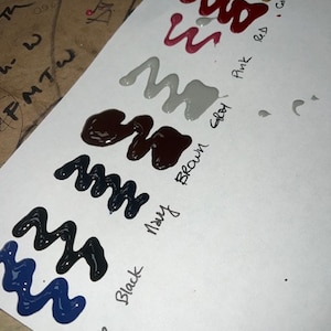 May include: A white sheet of paper with paint swatches in various colours, including blue, black, brown, grey, pink, red, and camel. The colours are labelled with their names in black ink.