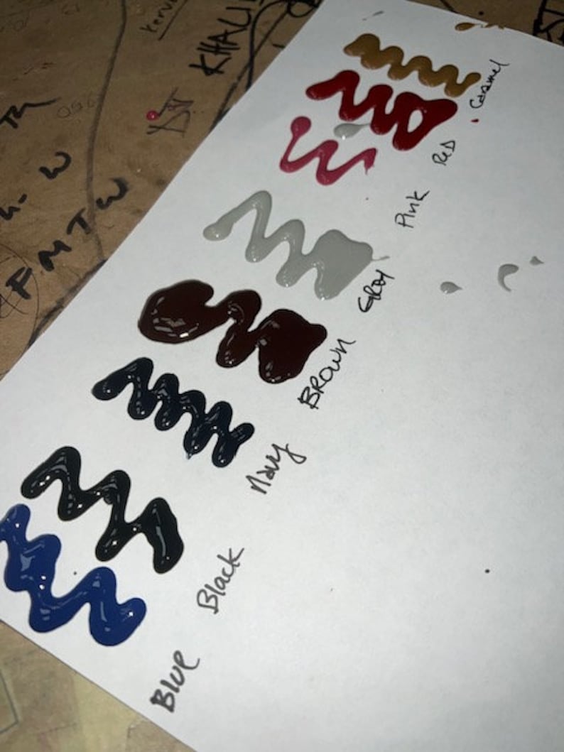 May include: A white sheet of paper with paint swatches in various colours, including blue, black, brown, grey, pink, red, and camel. The colours are labelled with their names in black ink.