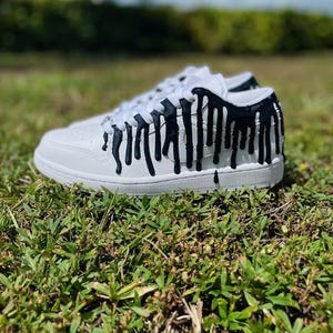 May include: White trainers with black paint dripping down the sides. The paint looks like it is melting down the shoe.