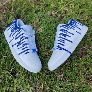 May include: White trainers with blue paint dripping down the sides. The trainers have a white sole and laces.