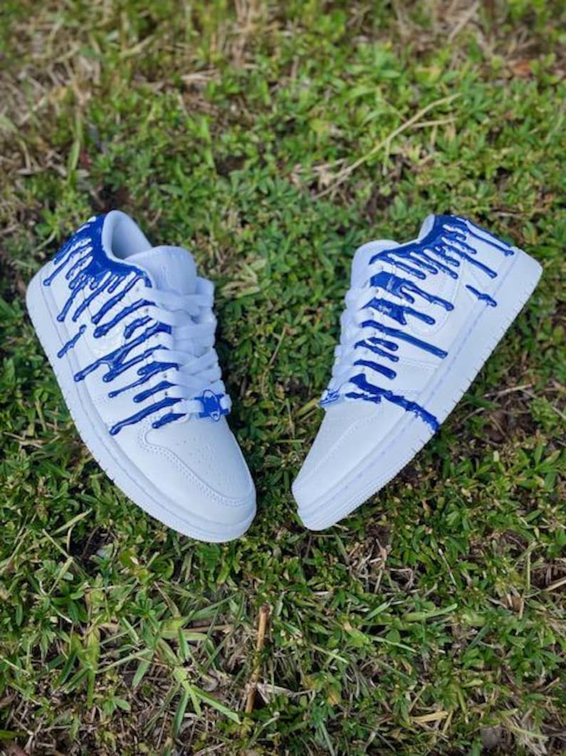 May include: White trainers with blue paint dripping down the sides. The trainers have a white sole and laces.