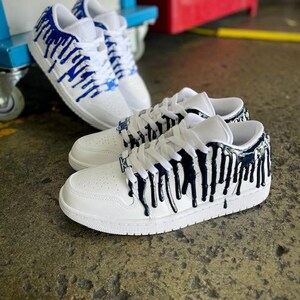 May include: Two pairs of white trainers with blue and black paint dripping down the sides. The trainers have a white sole and laces.