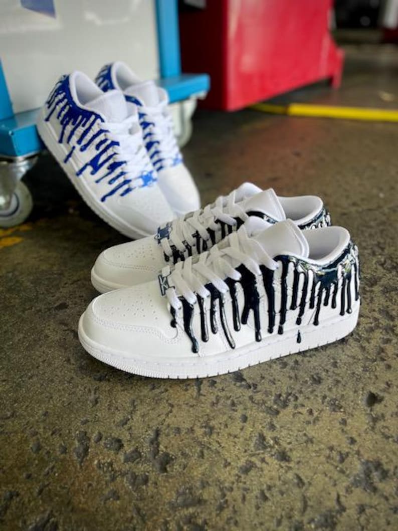 May include: Two pairs of white trainers with blue and black paint dripping down the sides. The trainers have a white sole and laces.