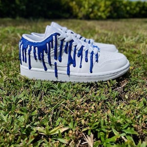 May include: White low-top trainers with blue paint dripping down the sides. The trainers have a white sole and white laces.