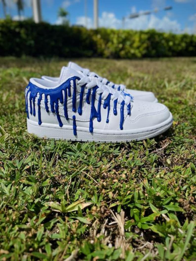 May include: White low-top trainers with blue paint dripping down the sides. The trainers have a white sole and white laces.