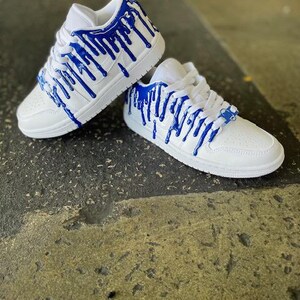 May include: White trainers with blue paint dripping down the sides. The trainers have a white sole and a white upper with blue paint dripping down the sides.