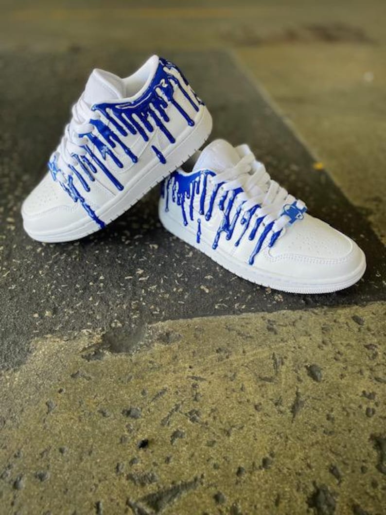 May include: White trainers with blue paint dripping down the sides. The trainers have a white sole and a white upper with blue paint dripping down the sides.