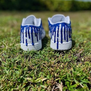 May include: A pair of white trainers with blue paint dripping down the sides. The trainers are on a green grassy surface.