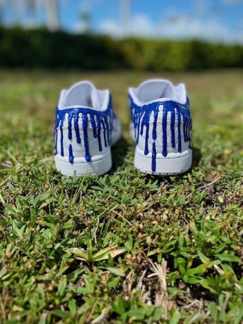 May include: A pair of white trainers with blue paint dripping down the sides. The trainers are on a green grassy surface.