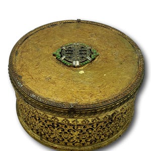Op de afbeelding: Een ronde, antieke goudkleurige doos met een decoratieve filigraanbasis en deksel. Het deksel heeft een centrale versiering met groene en heldere accenten. De doos heeft een vintage uitstraling.