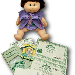 May include: A Cabbage Patch Kids doll with brown hair and a purple outfit. The doll is surrounded by birth certificates, adoption papers, and a small book. The doll's outfit includes a purple jacket and a floral print dress.