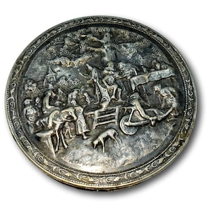 May include: A round, silver-toned metal plate with a detailed relief scene of a village gathering. The plate features figures, animals, and architectural elements within a decorative border. The relief work is intricate, showcasing a historical or pastoral theme.